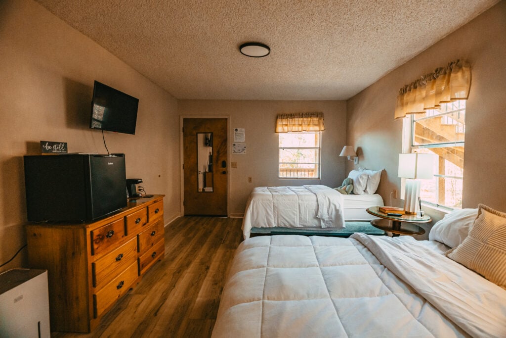 Guest room at The Bridges in Wayne County with two beds, wood flooring, large windows, dresser, mini fridge, coffee station, and TV.