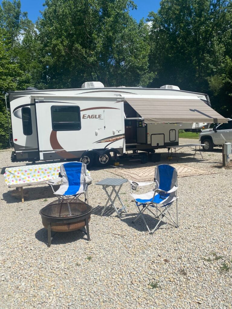 White travel trailer with extended awning parked on gravel, surrounded by blue camping chairs and a metal fire pit. Trees loom in the background.