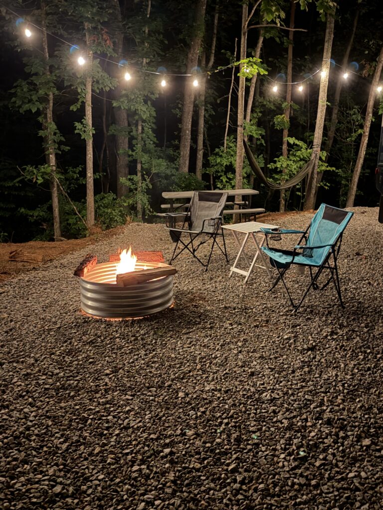 Night camping scene: string lights above a gravel clearing, a glowing fire pit, two camping chairs and a small table with a hammock in the trees nearby.