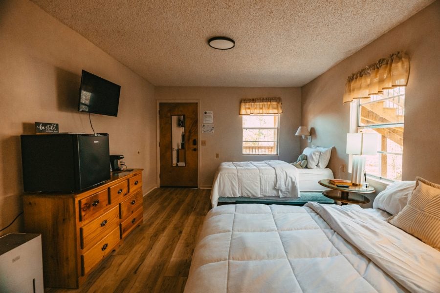 Guest room at The Bridges in Wayne County with two beds, wood flooring, large windows, dresser, mini fridge, coffee station, and TV.
